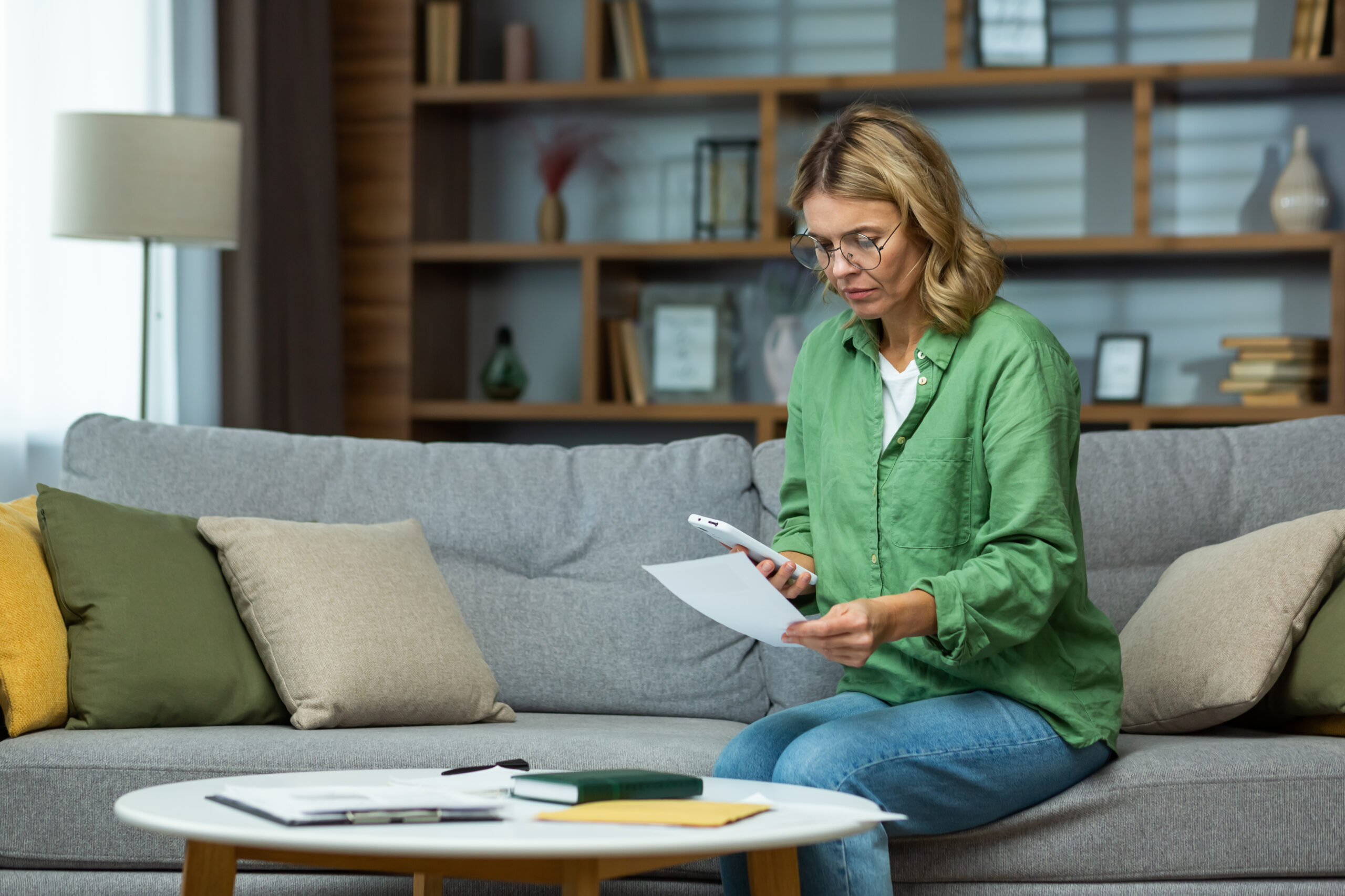 Woman sitting on a sofa, reviewing documents and holding a smartphone.