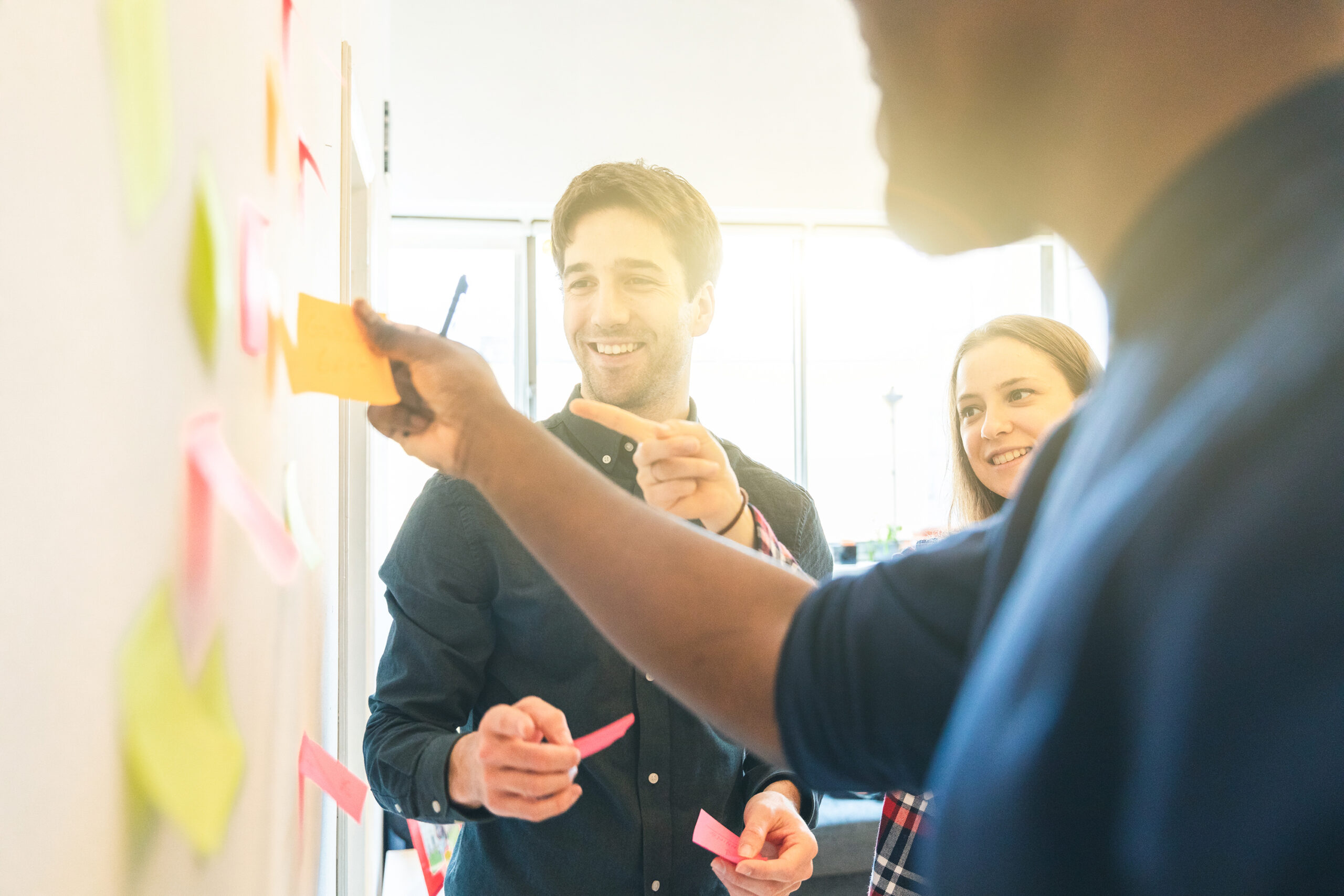 A group of smiling colleagues engages in a brainstorming session, placing sticky notes on a board in a bright, collaborative workspace