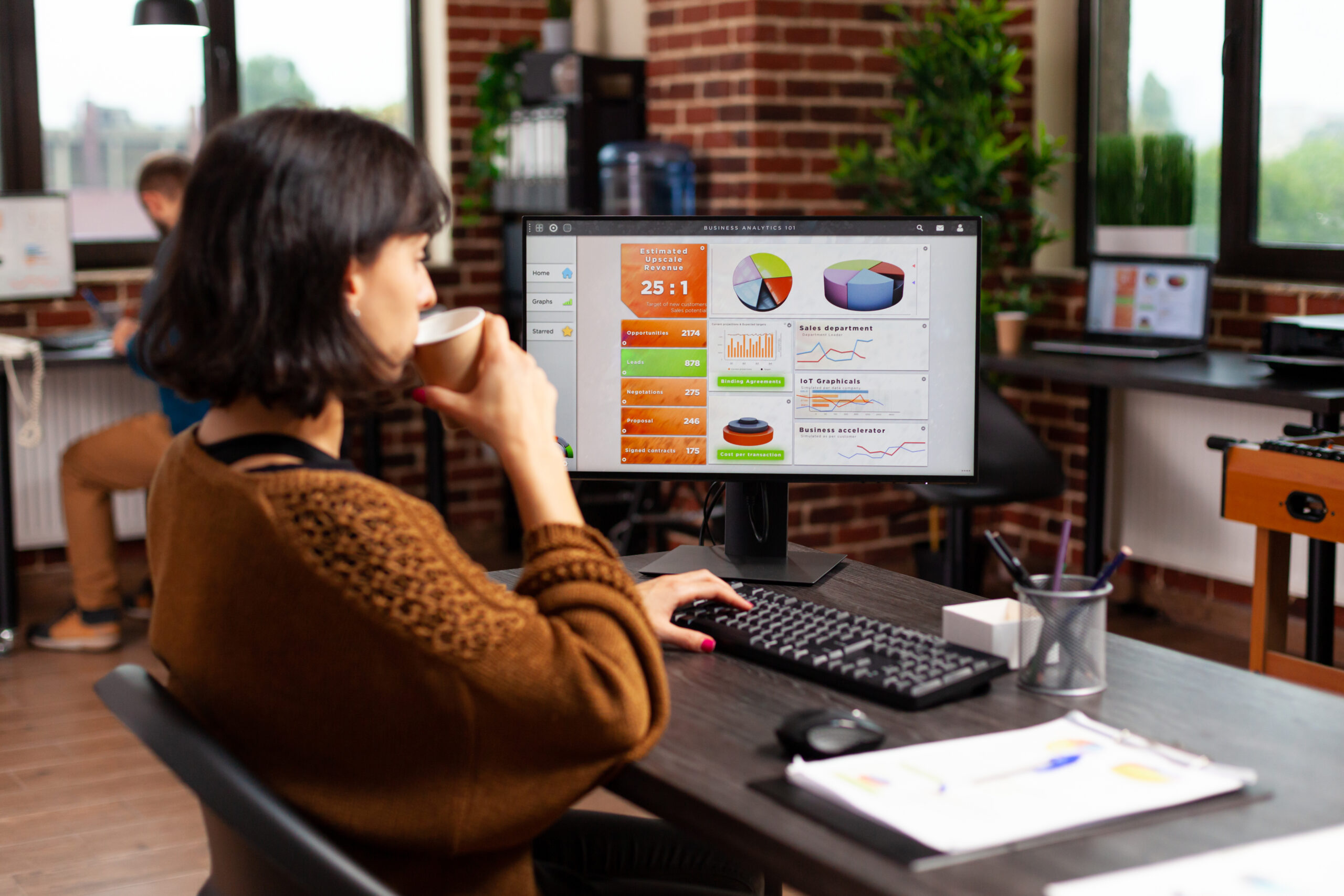 A businesswoman sits at her desk, analyzing data on her computer screen while holding a cup of coffee in a modern office environment