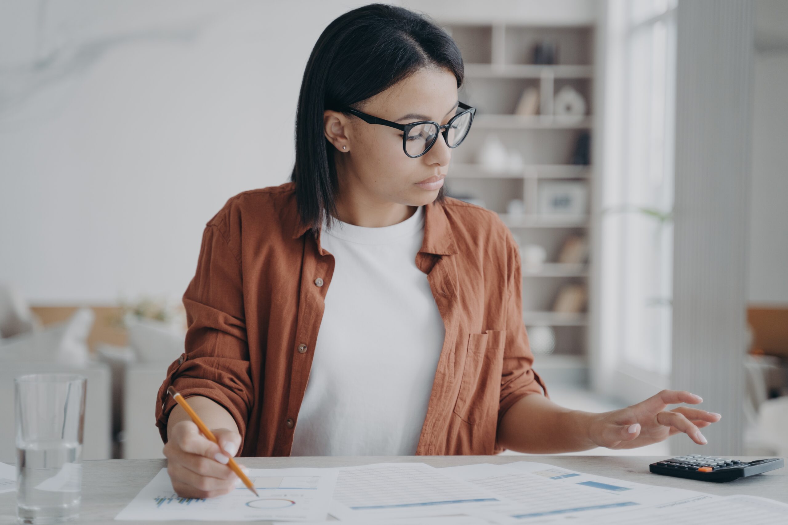 Businesswoman wearing glasses counts expenses on calculator, planning company budget, working with papers. Focused female accountant freelancer works on report. Financial management.