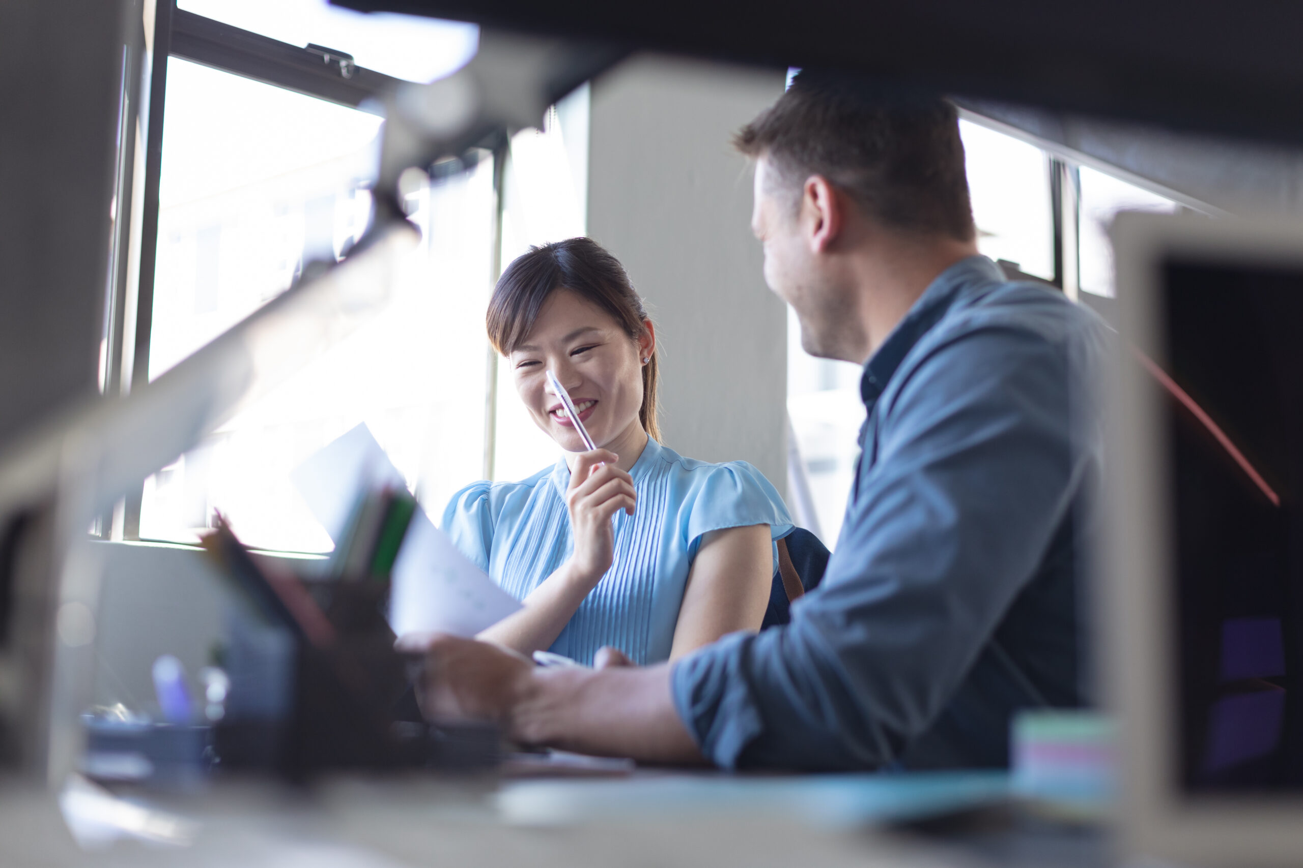 Low angle close up of a Caucasian male and an Asian female business creative working together and smiling in a casual modern office, with blurred euipment on their desk in the foreground