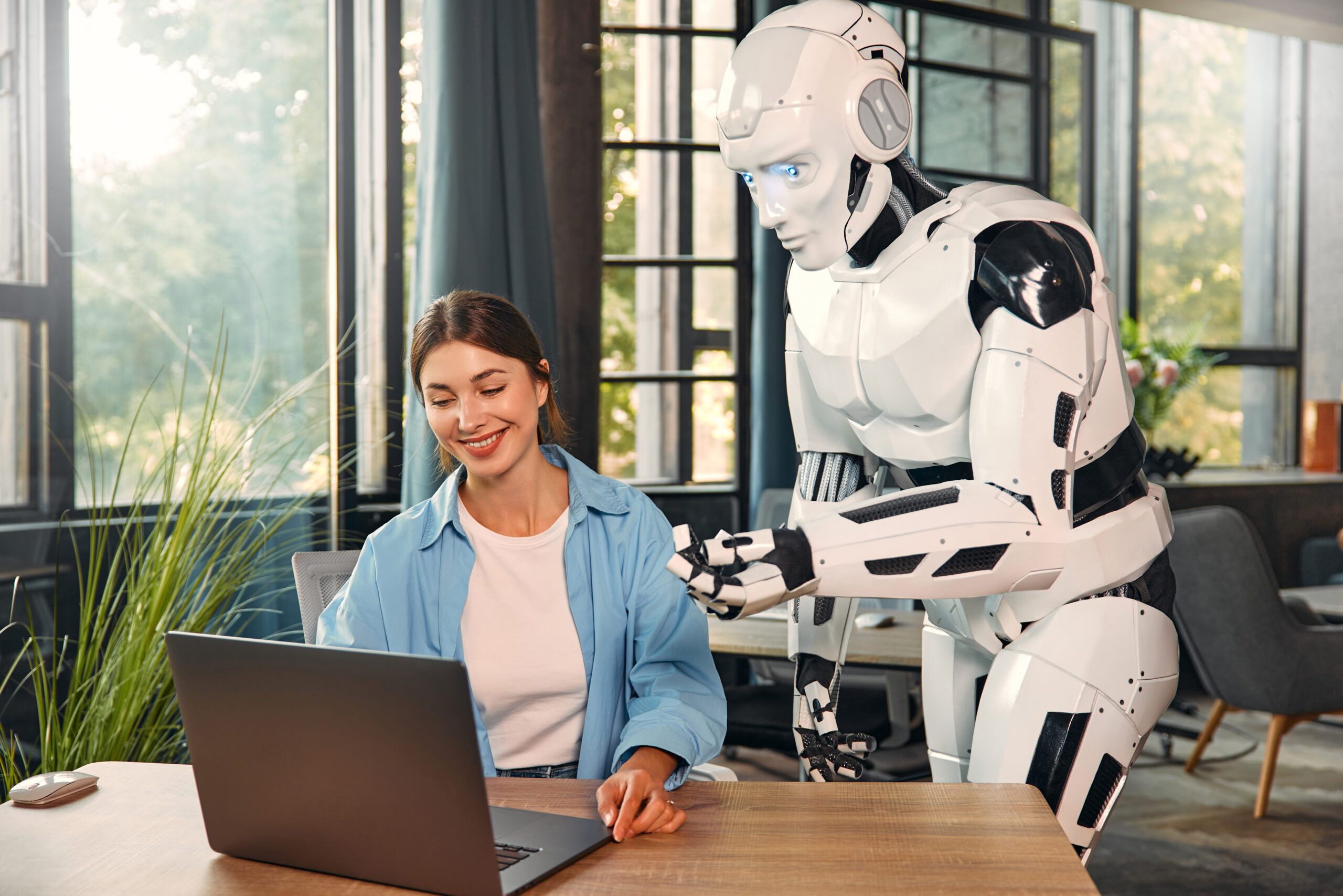 Robot and woman working on laptop in office Young woman and a humanoid robot working while sitting at a laptop in a modern office. Collaboration between humans and artificial intelligence.