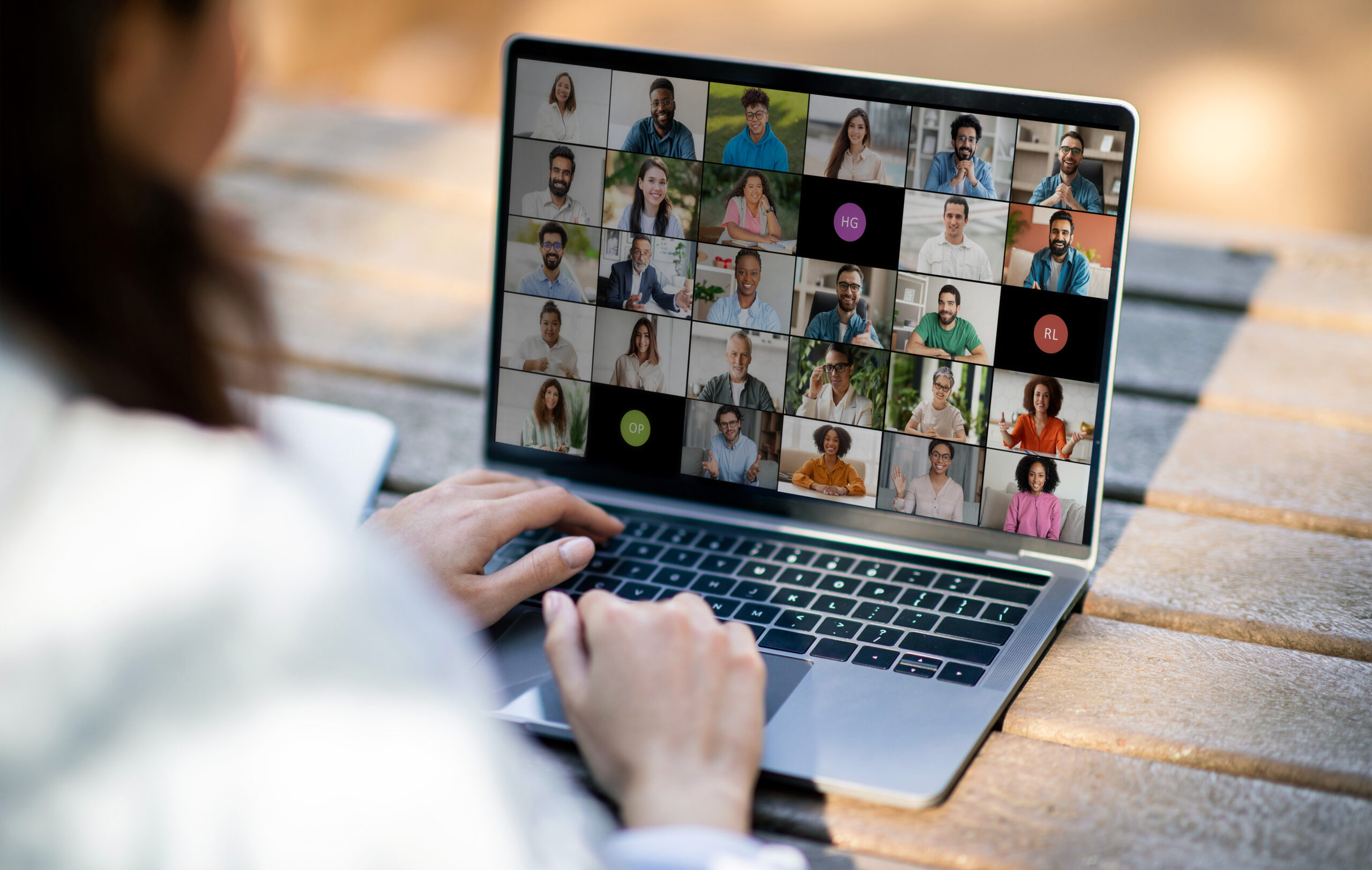 Lady Participating in Virtual Business Meeting Outdoors During Daytime Lady sitting at an outdoor table, actively engaging in a virtual business meeting on a laptop. The screen displays numerous participants in different video windows.